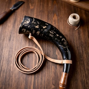 May include: A black drinking horn with a light brown leather strap and a wooden base. The horn has a curved shape and a textured surface. The strap is coiled. A knife and twine are in the background.