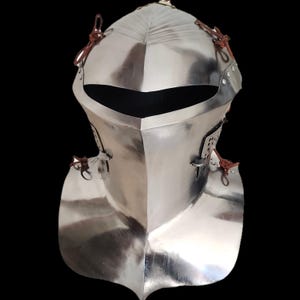 Op de afbeelding: Een gepolijste, zilverkleurige middeleeuwse helm met een puntig vizier en een nekbeschermer. De helm heeft klinknagels en leren riemen voor bevestiging. Het ontwerp is reflecterend, met een donkere achtergrond.