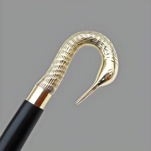 May include: A walking cane with a gold-colored bird head handle and a black wooden shaft. The bird's head is detailed with textured patterns and a smooth, polished beak. The cane has a gold-colored metal band at the top.