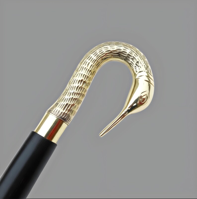 May include: A walking cane with a gold-colored bird head handle and a black wooden shaft. The bird's head is detailed with textured patterns and a smooth, polished beak. The cane has a gold-colored metal band at the top.