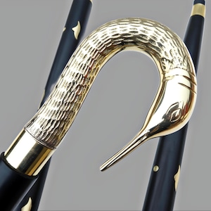 May include: A decorative walking cane with a gold-colored swan head handle. The handle has a textured finish and a smooth, polished beak. The black shaft is accented with gold-colored details.