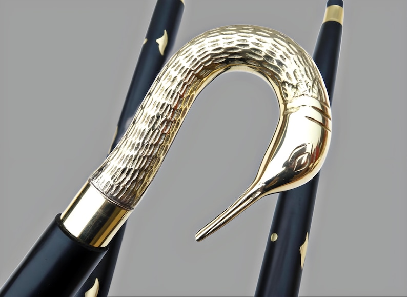 May include: A decorative walking cane with a gold-colored swan head handle. The handle has a textured finish and a smooth, polished beak. The black shaft is accented with gold-colored details.