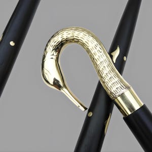May include: A black walking cane with a gold-colored swan head handle. The cane's shaft has gold accents. The swan's head is intricately detailed, and the cane appears to be made of wood and metal.