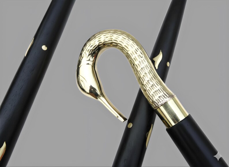 May include: A black walking cane with a gold-colored swan head handle. The cane's shaft has gold accents. The swan's head is intricately detailed, and the cane appears to be made of wood and metal.