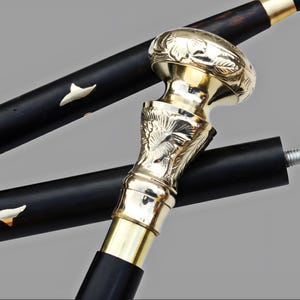 May include: A close-up of a black walking cane with a decorative gold-coloured handle. The handle has an ornate floral design. The cane's shaft has white decorative accents. The cane is a classic accessory.