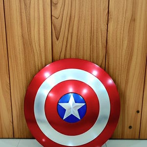 May include: A red, white, and blue Captain America shield. The shield is round with a silver outer ring, a red center, and a blue circle with a white star in the middle. The shield is a prop or collectible.