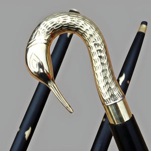 May include: A decorative walking cane with a gold-colored bird head handle and a black shaft. The bird head has a textured surface. The shaft is black with gold accents. The cane is angled against a light gray background.