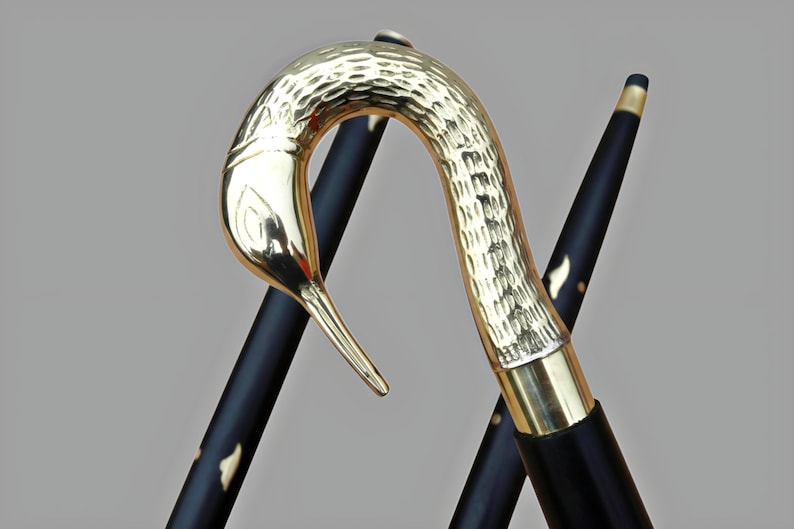 May include: A decorative walking cane with a gold-colored bird head handle and a black shaft. The bird head has a textured surface. The shaft is black with gold accents. The cane is angled against a light gray background.