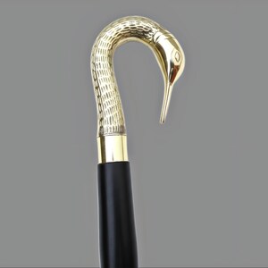 May include: A decorative walking cane with a bird-shaped handle. The handle is gold-colored and features intricate textured detailing. The shaft is black with a gold band at the top. The cane is designed for support and style.