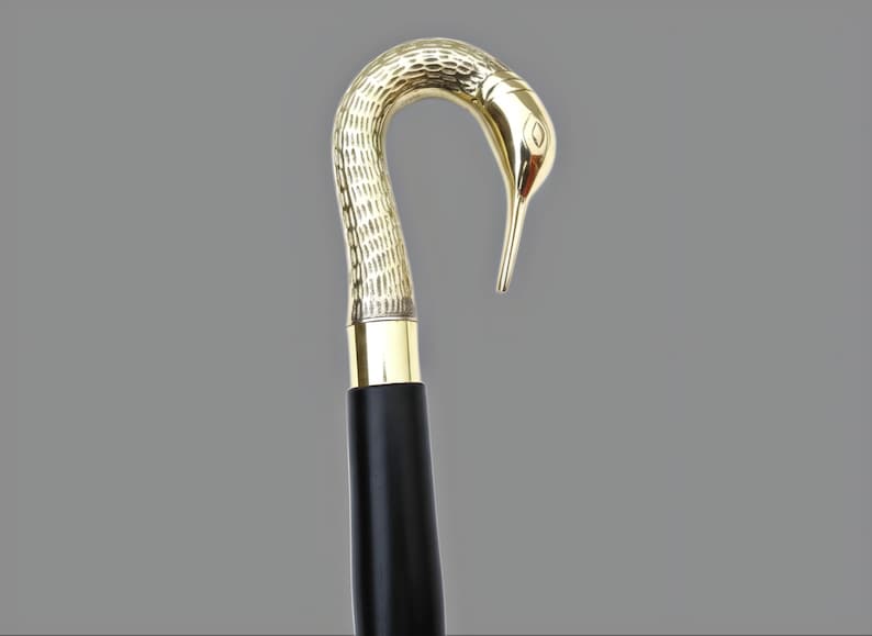 May include: A decorative walking cane with a bird-shaped handle. The handle is gold-colored and features intricate textured detailing. The shaft is black with a gold band at the top. The cane is designed for support and style.