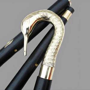 May include: A black wooden walking cane with a gold-colored bird-shaped handle. The handle has a textured surface, and the cane features gold accents. The cane is angled, showcasing its design and craftsmanship.