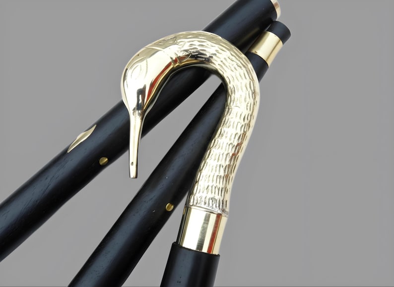 May include: A black wooden walking cane with a gold-colored bird-shaped handle. The handle has a textured surface, and the cane features gold accents. The cane is angled, showcasing its design and craftsmanship.
