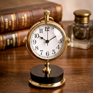 Könnte beinhalten: Eine runde Schreibtischuhr aus Messing mit weißem Zifferblatt, schwarzen arabischen Ziffern und schwarzen Zeigern. Die Uhr ist auf einem zylindrischen schwarzen Sockel mit Messingrand montiert. Ein kleiner Messingknopf befindet sich oben am Gehäuse, das über einen zentralen Messingfuß mit dem Sockel verbunden ist.