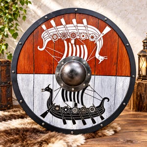 May include: A circular wooden shield featuring a black metal rim and a central steel boss. The upper half is stained brown with a white longship graphic, while the lower half is painted white with a black longship graphic and oars.
