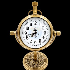 May include: A brass-colored nautical-themed clock with a white face and black numerals. The clock features an anchor design and is mounted on a decorative base. The clock is approximately 6 inches tall.