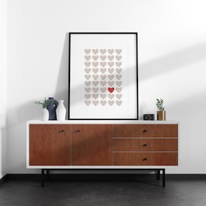 Puede incluir: Una impresión enmarcada con un patrón de corazones beige y un corazón rojo. La obra de arte está sobre un mueble blanco y de madera con patas negras. Jarrones decorativos y una pequeña planta están sobre el mueble. El mueble mide unos 150 cm de ancho.