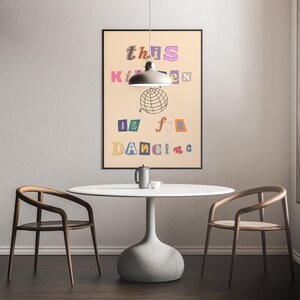 Puede incluir: Escena de comedor con una mesa redonda blanca y dos sillas de madera. Un póster enmarcado con el texto "this kitchen is for dancing" cuelga de la pared. Una tetera y una taza están sobre la mesa.