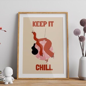 Puede incluir: Una impresión artística enmarcada con las palabras "KEEP IT CHILL" en rojo. La obra de arte presenta una figura con pantalones rojos y una parte superior negra colgando de una cuerda en un círculo rosa. También se ve un jarrón con flores de pompón moradas y una figura decorativa.