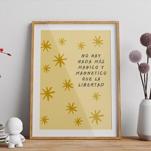 Puede incluir: Un cuadro enmarcado con fondo amarillo y diseños dorados de estrellas. El cuadro tiene el texto "NO HAY NADA MÁS MAGICO Y MAGNETICO QUE LA LIBERTAD". Los elementos decorativos incluyen un jarrón con ramas rojas y un jarrón blanco con tallos de pompones.