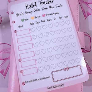 Könnte beinhalten: Ein rosa Gewohnheitstracker mit dem Text "Habit Tracker" und "You're Doing Better Than You Think." Der Tracker enthält Felder für Gewohnheiten, Wochentage und einen Abschnitt zum Aufschreiben, worauf man stolz ist. Der Tracker liegt auf einem rosa Planer.