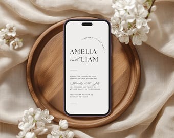 May include: A smartphone screen shows a wedding invitation with the names Amelia and Liam in black script. The phone is on a wooden tray, with white flowers and a beige fabric background. The invitation text includes the date.