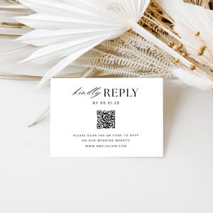 May include: A white RSVP card with the text "kindly REPLY" and a QR code. The card also includes the date "06.01.26" and website information. The card is placed on a white surface with dried floral accents.