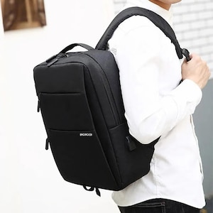 Könnte beinhalten: Ein schwarzer Rucksack mit rechteckiger Form und einem oberen Griff. Er hat eine Vordertasche und verstellbare Schultergurte. Der Markenname "BROMOGE" ist sichtbar. Der Rucksack wird getragen.