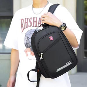 May include: Black backpack with a red and silver accent. The backpack has a zippered main compartment and a smaller front pocket. A red emblem is on the front. The backpack is being worn over the shoulder. The person is wearing a white t-shirt with the text "RHSHA".