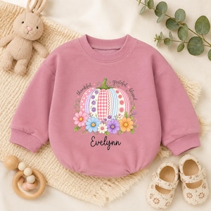 May include: A dusty pink long-sleeve sweatshirt featuring a central graphic of a pumpkin with striped and checkered patterns, surrounded by colourful flowers. The text "thankful" and "grateful blessed" surrounds the pumpkin, with the name "Evelynn" printed below.
