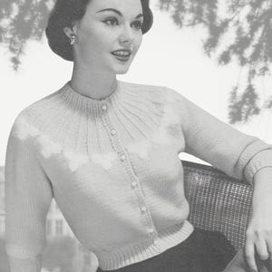 Könnte beinhalten: Eine helle, geknöpfte Strickjacke mit Dreiviertelärmeln, einem gerippten Rundhalsausschnitt und einem dekorativen Bogenmuster auf der Brust. Das Kleidungsstück wird mit einem dunklen Rock und einem kleinen, gefalteten Stoffaccessoire kombiniert.