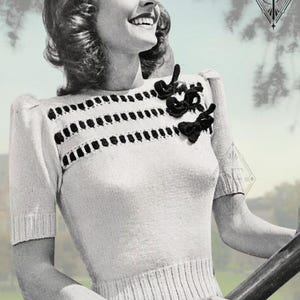 May include: A short-sleeved knit sweater in a light color featuring a ribbed waist and sleeve cuffs. The upper chest area displays horizontal rows of black rectangular cutouts and three decorative black fabric bows on the left side.