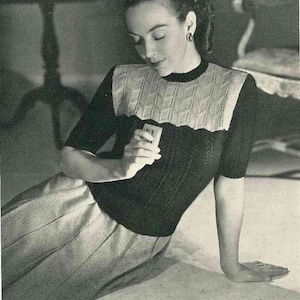 May include: A woman wearing a black and white knitted sweater with a skirt. She is holding a playing card in her hand.