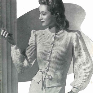 May include: A vintage black and white image of a woman wearing a white, open-weave cardigan with a button-up front and a tie at the waist. The cardigan has long, puffed sleeves. The text "DESIGN No. 2590" is in the upper right corner.