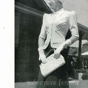 May include: A black and white photograph of a woman wearing a white jacket with a distinctive criss-cross design, a white turban, and a white handbag. She is standing in front of a wooden structure.