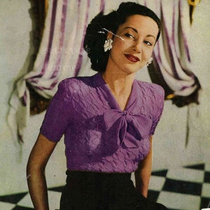 May include: A purple knitted sweater with a bow at the neckline. The sweater has a cable knit pattern and short sleeves.