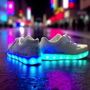 May include: White LED light-up trainers with the words "Just Married" and "Mrs. Givens" inscribed. The shoes have blue and green lights around the soles, with the date "6-10-25" on one shoe. The background is blurred with city lights.