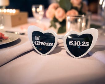 Op de afbeelding: Witte hartvormige zonnebril met zwarte glazen, met de tekst "THE Givens" op het linkerglaasje en "6.10.25" op het rechterglaasje. De zonnebril ligt op een lichtroze tafelkleed, wat duidt op een bruiloft of evenement.