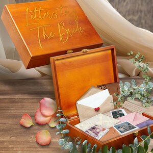 May include: Wooden keepsake box with the words "Letters to The Bride" in gold script. The box is open, revealing letters, photos, and a small red heart. Rose petals and greenery are scattered around the box.