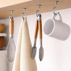 Op de afbeelding: Keukengerei hangend aan haken onder een houten plank. Een garde, een beige handdoek, een tang en een witte mok hangen. De garde en tang hebben houten handvatten. De handdoek heeft een wafelweefsel.