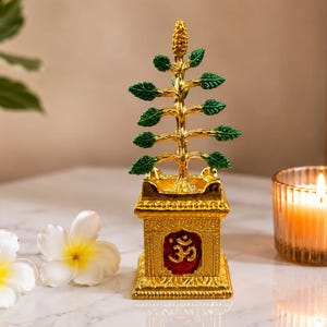 May include: A gold-colored metal sculpture of a tree with green leaves, mounted on a square base featuring a red inset panel with a gold Om symbol. The tree has a textured trunk and branches, with a pointed top, standing upright on a flat surface.