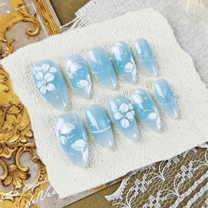 May include: A set of ten pale blue false nails, each adorned with white floral patterns. The oval-shaped nails are displayed on a white fabric. The background features a decorative card and lace trim.
