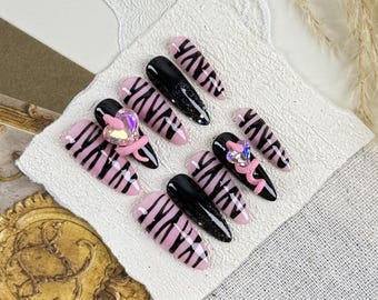 Uñas postizas con diseño de rayas de cebra en rosa y negro, con elegantes acabados almendrados, diamantes de imitación y lazos, ideales para looks atrevidos en citas, fiestas y mujeres seguras de sí mismas.