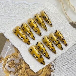 Könnte beinhalten: Ein Satz von zehn langen, mandelförmigen Kunstnägeln. Jeder Nagel ist mit einer goldenen Basis und schwarzen Leopardenflecken bemalt. Die Nägel sind auf einer weißen, strukturierten Oberfläche angeordnet.