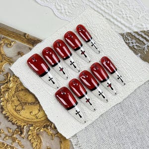 May include: A set of ten artificial nails. Each nail is painted a deep, glossy red with a black tip and a small cross design. The nails are arranged on a white surface, with decorative elements in the background.