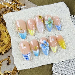 May include: A collection of ten artificial nails, each with a nude base and diverse decorations. Some nails have blue and yellow tips, while others feature gold accents, pearls, and floral designs. The nails are displayed on a textured white surface.