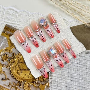 May include: A collection of ten artificial nails. They feature a nude to red gradient, leopard print detailing, and are adorned with rhinestones and a large, iridescent gem. The nails are designed for cosmetic use.