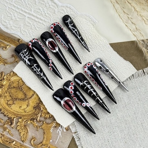 Könnte beinhalten: Ein Set aus zehn langen, spitzen, schwarzen Kunstnägeln. Jeder Nagel hat einzigartige Designs, darunter rote und weiße Akzente, silberne Verzierungen und einen zentralen roten Edelstein. Die Nägel sind auf einem weißen Tuch angeordnet.