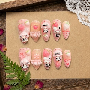 May include: A set of ten pink press-on nails with various designs. Each nail features a different design, including strawberries, bunnies, and bows. Some nails have 3D embellishments. The nails are arranged on a brown surface.