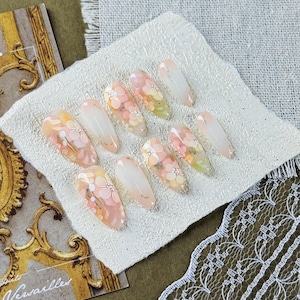 Puede incluir: Un conjunto de diez uñas artificiales con un diseño floral. Las uñas, de forma ovalada, presentan un degradado de colores melocotón, amarillo y blanco. Cada uña está adornada con delicados patrones florales y detalles de purpurina plateada.