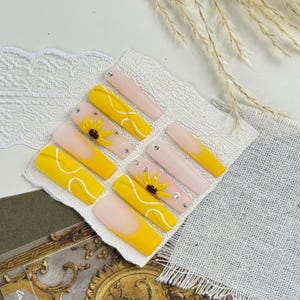 May include: A set of press-on nails featuring a mix of designs. Some nails are yellow with white swirls, others are pale pink with yellow tips. Several nails have small sunflower accents and rhinestones.